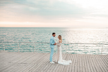 Fototapeta premium Young bride, fashionable, blonde model in luxury wedding dress with groom on the coast. Couple walks at the pier on sunrise