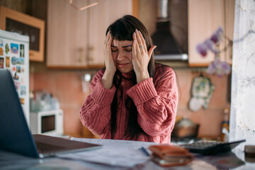 Sad emotional woman holding her head over utility bills in front of a laptop. The concept of rising...