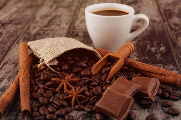 cup of coffee with chocolate, cinnamon sticks,   and coffee beans on wooden background