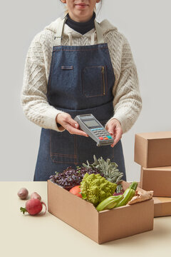 Woman in apron holding terminal by box with vegetables