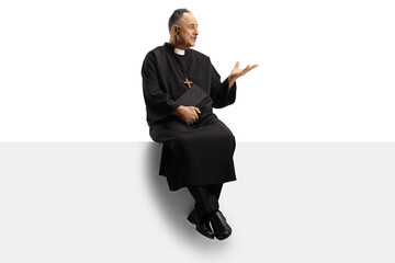 Full length shot of a mature priest sitting on a blank panel and gesturing with hand