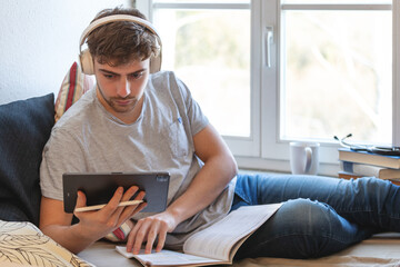 Junger Medizinstudent studiert online von zu Hause aus. Er liegt auf seinem Bett vor einem Fenster, trägt Kopfhörer, blickt konzentriert in sein Tablet und blättert in Unterlagen - obrazy, fototapety, plakaty