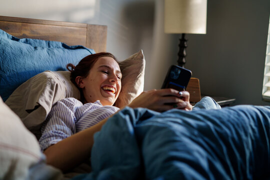A young woman smiles in bed browsing her phone - Powered by Adobe