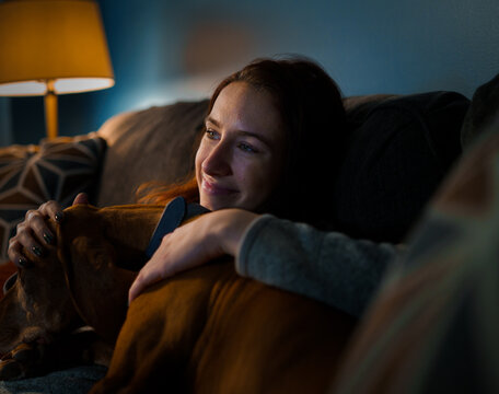 A young white woman relaxes on the couch with her dog