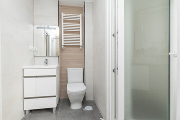 Tiled toilet with large white tiles, white porcelain sink with glass mirror and white wooden cabinet and white aluminum towel radiator