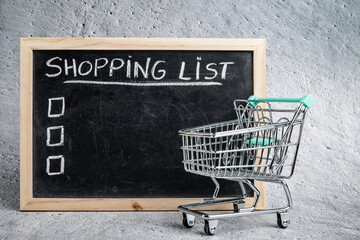 Empty shopping cart next to slate with wooden frame. Cement background. Slate with shopping list. Items to mark with check list. Copy space to put personalized items.