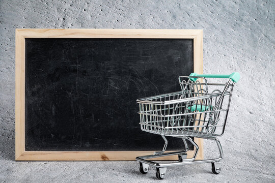 Empty shopping cart next to slate with wooden frame. Cement background. Copy space on the blackboard on the left side.