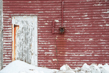 A white faded wood single door in a red vintage wooden barn. The hinges on the door are rusty and there's stain running down the paint peeling boards. There's white snow in front of the shed.  