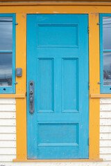 A colorful teal blue color wooden panel door with yellow decorative trim on a white exterior wall. There's a long brass door handle on the wood door and a glass window on both sides of the door. 