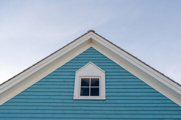 An upward view of a vintage blue wooden building with thick white trim, a small double hung window, with four panes of glass and a frame shingled roof. The roof has white trim and black shingles.