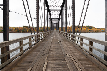 Steel trestle bridge with wooden deck over a large river in Newfoundland.  The bridge is for foot traffic and ATV usage. The sky is clear blue and land and houses can be seen in the background