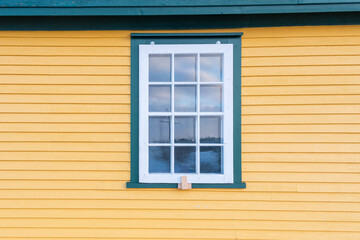 The exterior of a vintage yellow and green colored exterior wall with narrow wood cape cod clapboard siding. In the center of the house, there's a closed single hung window with a mountain reflection.