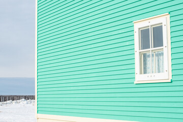 A traditional wooden clapboard wall of a vibrant teal green house with a single hung window with cream-colored trim. There's a cloudy background, snow, seawall, and a blue ocean in the background. 