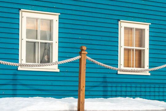 The exterior wall of a blue wooden clapboard style house with two single hung windows. There's white trim around the old windows, a rope fence in front of the building, and snow on the ground. 