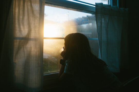 Young girls looks out window with sun setting outside 