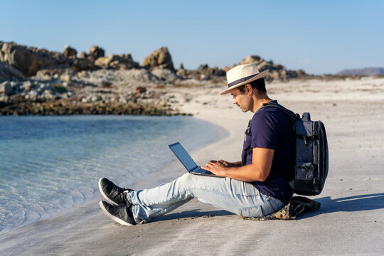 latin man sitting on the beach with laptop alone doing telecommuting or remote work