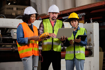 Safety uniform workers and industrial engineers with hardhat use laptop computer to check and control machines. Three professionals work in paper manufacturing factory, maintain production equipment.