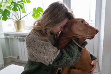 Middle age woman hugging her beloved Wirehaired Vizsla dog at home. Female owner kissing and...