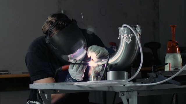 A fabricator TIG welding stainless steel exhaust pipe with pie cuts. 
Static shot from far away.