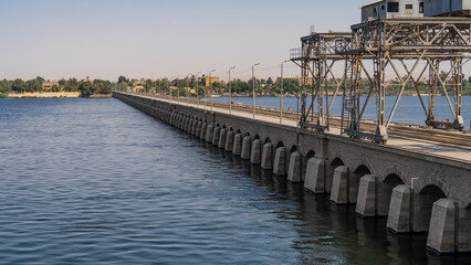 Concrete dam of the Nile sluice system in Esna. A pedestrian zone, technological metal structures are visible. On the far shore - green vegetation, houses. Blue water, clear sky. Egypt