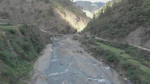 Rocky river bed excavator pulling river rock for cement production bridge spanning over water in valley connecting communities in Kabayan Benguet Philippines wide slow approaching aerial