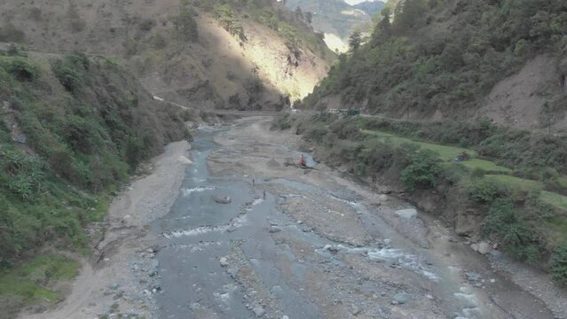 Rocky river bed excavator pulling river rock for cement production bridge spanning over water in valley connecting communities in Kabayan Benguet Philippines reverse reveal ascending aerial