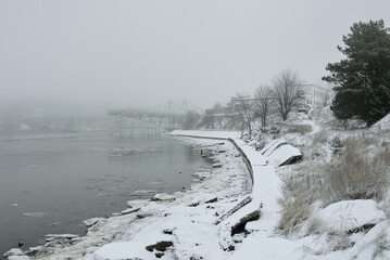 Frozen river, swing bridge, embankment, city park. Snow, thick fog. Concept winter urban landscape. Architecture, logistics, infrastructure themes - 493394083