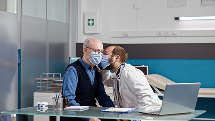 Doctor consulting retired patient with professional otoscope, doing ear checkup examination in medical cabinet. Elder man and male physician with face mask doing otology consultation.