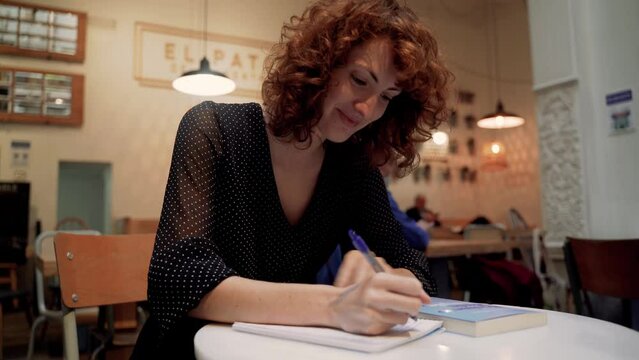 Educated Young Woman Writing Notes On Paper Sitting At Cafe Table