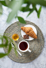Breakfast concept with a cup of tea , honey and a croissant on a marble table and green branches close-up. Top view. Vertical orientation
