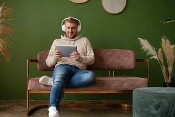 Caucasian blond young adult man with headphones,sitting on sofa at home working with tablet computer in modern beautiful interior indoors.Work,business from home,surfing internet,listening to podcast
