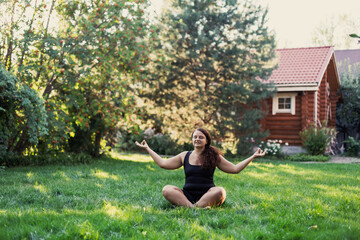 Young over-weighted woman doing yoga sitting in lotus position outside on backyard of cottage with trees and wooden house in background. Body positive. Equality. 