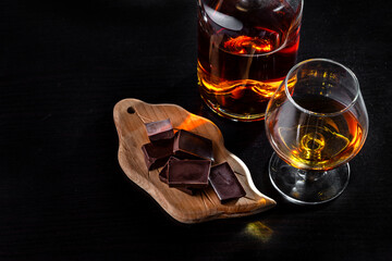 A glass of cognac, cognac in a bottle and pieces of chocolate on a dark background. Copy space