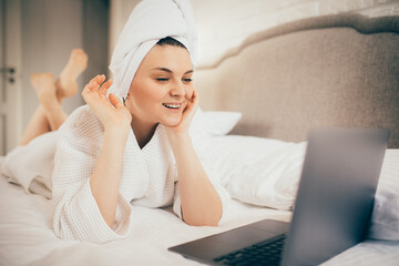 Young woman making video call from home using her laptop, lying in bed in white bathrobe and towel on head.