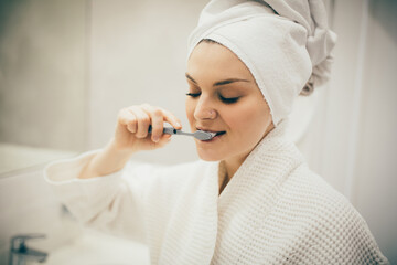 Young woman wearing white bathrobe and tower on head brushing her teeth using toothbrush and paste.