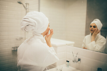 Fototapeta na wymiar Young woman wearing white bathrobe and towel on head applying cream on face. Daily beauty routine.