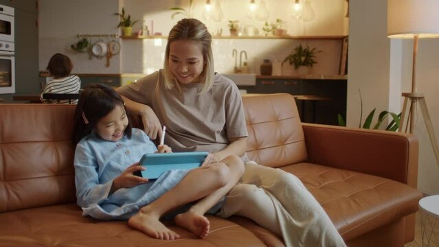 Happy Asian Family Using Tablet Computer With Children Mother Watching Daughter Play Game on Touchscreen Technology Sitting on Sofa. Using Education Application for Easy and Funny Learning