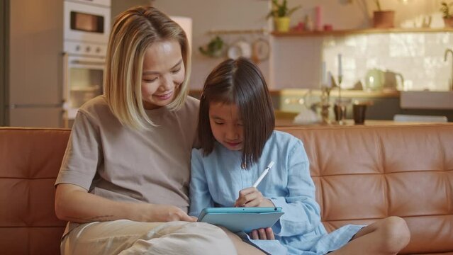 Family Concept. Mother and Daughter Play on Digital Tablet, Home With Tablet Device Draw With Pencel at Home, Doing Homework. Little Girl Smile Have Fun With Tutor Mother. Two Person Sit at Sofa