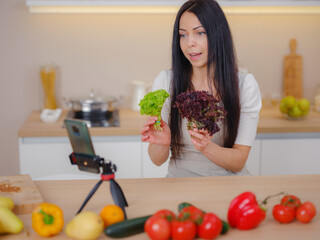 Professional beautiful happy young woman is blogging for her kitchen channel about healthy living in kitchen of her home and looking on smartphone on tripod