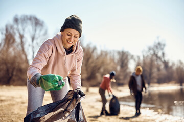 Happy woman picking up plastic bottles while cleaning beach with her friends.