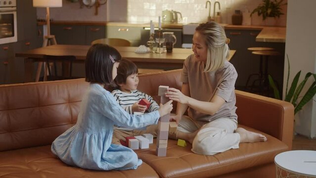 Young Asian Mother and Little Siblings Spend Time at Modern Flat Play Together Seated on Sofa, Enjoy Playtime Use Wooden Blocks. Babysitting Work, Leisure at Home with Kids Concept