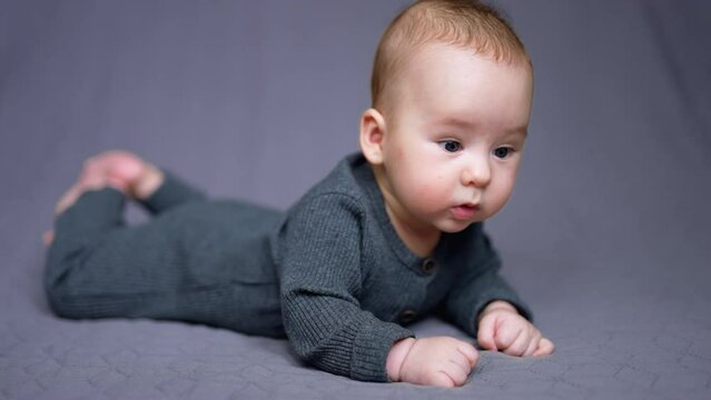 Sweet Caucasian child in grey outfit lies on belly on the bed. Beautiful child looking around with surprise. Grey backdrop.