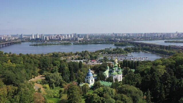 Capital of Ukraine - Kyiv city. Top view of cityscape, blue Dnipro, traffic on bridge over river and beautiful huge green trees in foreground. Real time full hd video footage.