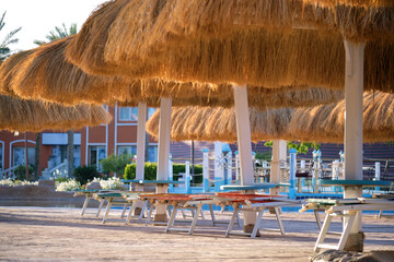 Naklejka na ściany i meble Empty deck chairs under straw shade umbrellas on swimming pool side in tropical resort. Summer vacations and getaway concept