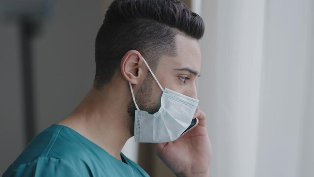 Latin man medical assistant doctor in medical mask consulting patient remote discussing issues by phone hispanic adviser using mobile healthcare app standing near window distant consultation concept