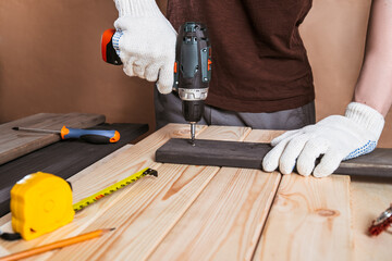 Close up portrait and details of caucasian male worker using electric screwdriver instrument in hand and repairing new wooden desk, home improvement concept.