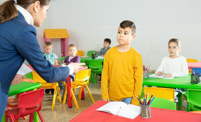 Upset boy listening reprimanding of strict female teacher during lesson in primary school