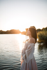A young pregnant woman in a chiffon dress stands on a pier by the river against the backdrop of an...