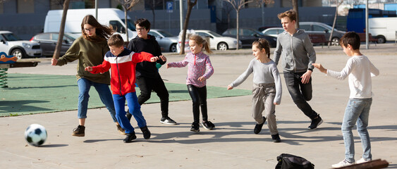 Large group of playful children running and kicking ball together at urban landscape