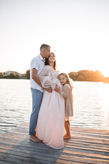 Fototapeta premium A husband with a pregnant wife and daughter is resting on the river bank, standing on the pier in summer sunny weather at sunset. couple in love man and woman hugging holding hands. A happy family